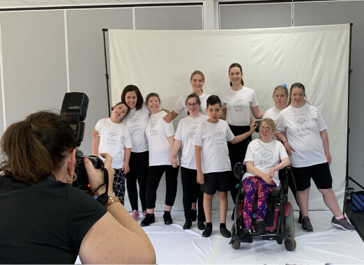 A group of All Abilties Australia students and their teachers, Miss Annabelle, Miss Georgia and Miss Catherina are showing their incredible diversity by all posing together in a group behind a white photo seet with a photographer taking their photo. They are all smiling proudly
