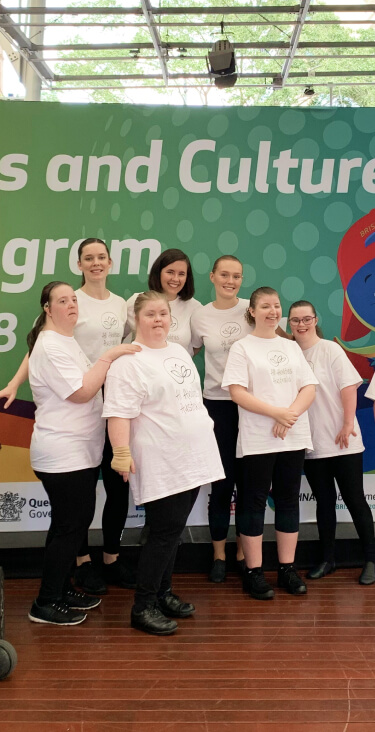 A group of students and their teachers are posing proudly behind the Arts and Culture Program 2020 poster that they performed at in on the stage in Queen St Mall